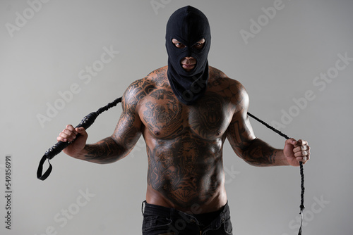 Muscular shirtless young man with whip in a mask.Brutal handsome man with tattooed body. Men tattoo casual fashion. Portrait of handsome male model. Muscular athletic sexy male with naked torso. photo