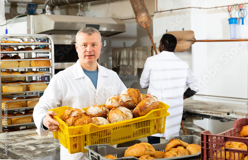 Portrait of professional baker working in bakehouse, carrying box with baked bread..