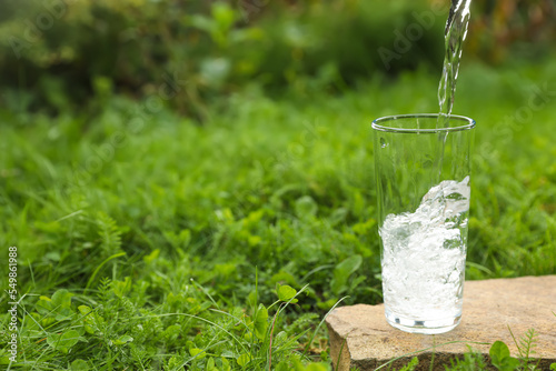 Pouring fresh water into glass on stone in green grass outdoors. Space for text