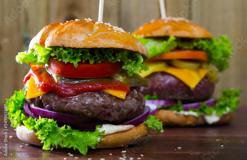 Two delicious burgers with cheese, tomato, pickled cucumbers, onion and lettuce on wood background