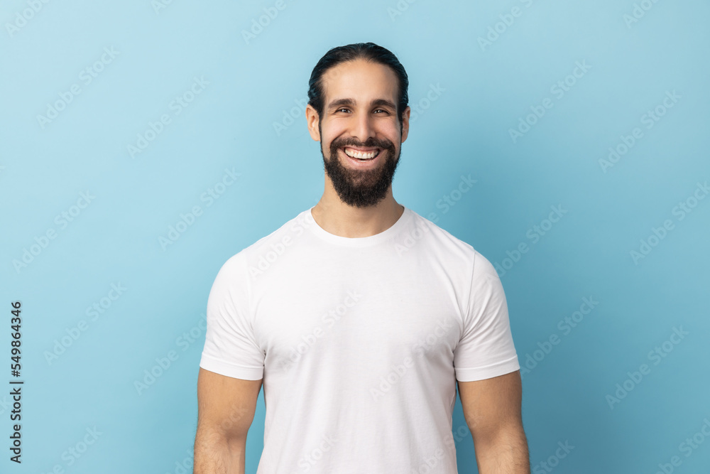 Portrait of bearded handsome man wearing white T-shirt standing looking at camera with satisfied face and smiling, expressing happiness. Indoor studio shot isolated on blue background. Portrait of bearded handsome man wearing white T-shirt standing looking at camera with satisfied face and smiling, expressing happiness. Indoor studio shot isolated on blue background.