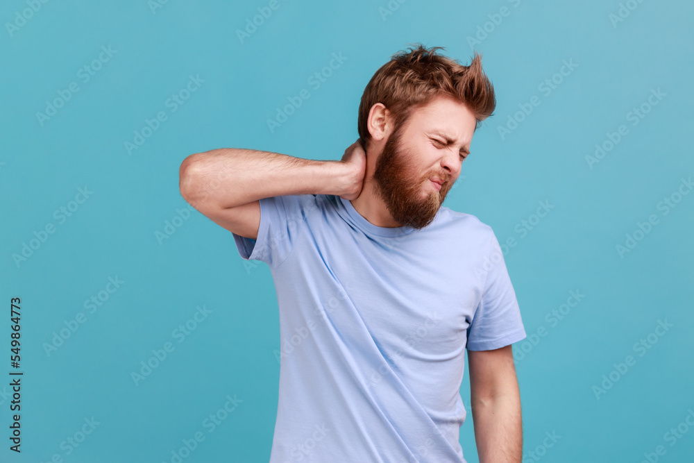 Portrait of unhealthy exhausted bearded man massaging aching neck with expression of unbearable suffer, having spine problems, back pain. Indoor studio shot isolated on blue background. Portrait of unhealthy exhausted bearded man massaging aching neck with expression of unbearable suffer, having spine problems, back pain. Indoor studio shot isolated on blue background.