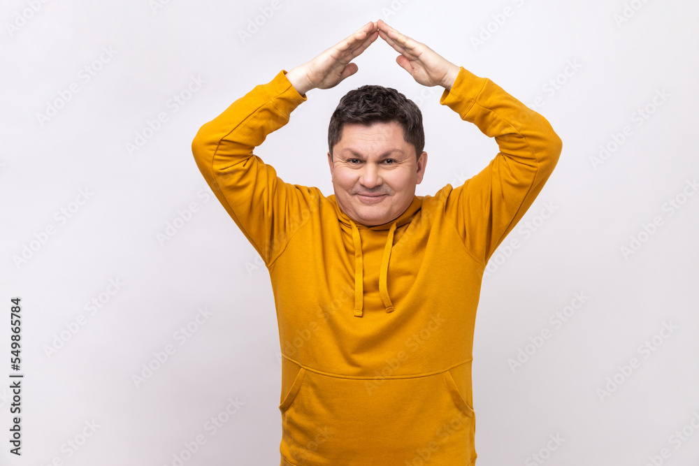 Portrait of cheerful middle aged man standing under roof house gesture and smiling, dreaming of real estate purchase, wearing urban style hoodie. Indoor studio shot isolated on white background. Portrait of cheerful middle aged man standing under roof house gesture and smiling, dreaming of real estate purchase, wearing urban style hoodie. Indoor studio shot isolated on white background.