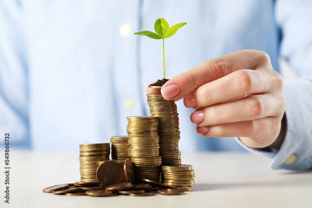 Woman putting coin with green sprout onto stack at white table, closeup. Investment concept Woman putting coin with green sprout onto stack at white table, closeup. Investment concept