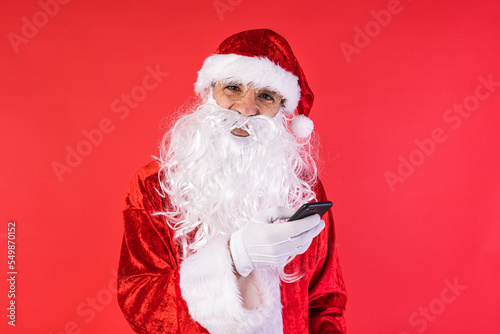 Man dressed as Santa Claus, consulting his mobile phone, on red background. Christmas, celebration, gifts, consumerism and happiness concept.