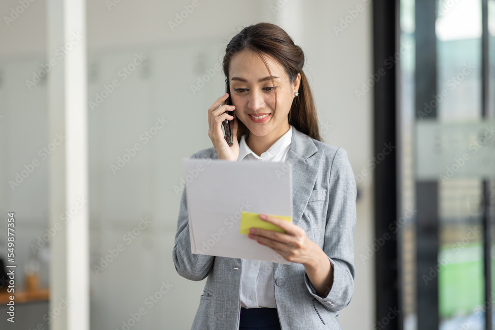 Young smiling Business asian woman talking on mobile phone, standing in workplace office with document in her hand Young smiling Business asian woman talking on mobile phone, standing in workplace office with document in her hand