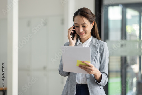 Young smiling Business asian woman talking on mobile phone, standing in workplace office with document in her hand