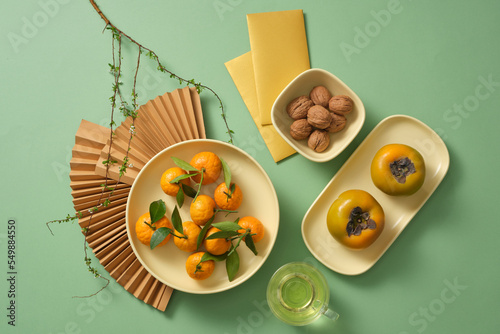 Set up New year table with fruits, nut and tea. Paper fans, lucky money, branches flower on green background. New year decoration idea. 