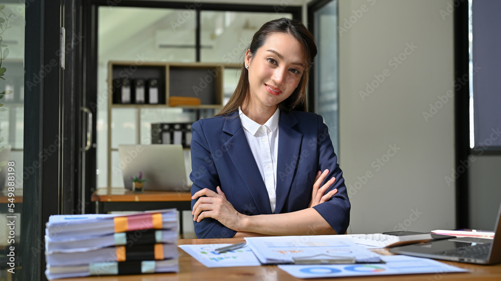 Attractive Asian businesswoman sitting at her desk, arms crosses, smiling and looking at camera. Attractive Asian businesswoman sitting at her desk, arms crosses, smiling and looking at camera.
