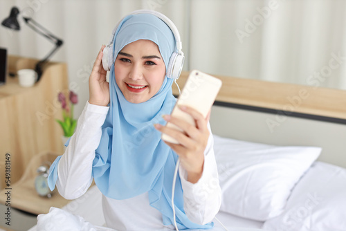 Portrait of beautiful asian female in muslim sleepwear watching online story on mobile phone, sit on bed and connected with wireless internet. Young cute girl with hijab listen music from smartphone