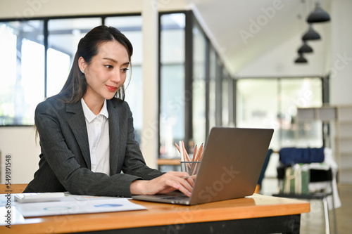 Professional Asian female accountant using laptop, typing on keyboard, working in her office.