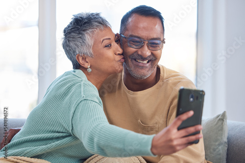 Phone, selfie and kiss with a senior couple posing for a picture in the living room of their home together. Smartphone, kissing and social media with a mature man and woman taking a photograph