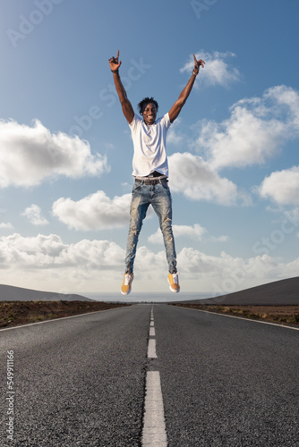 Cheerful positive black man enjoying life jumping in the air with raised fists over road in a desert landscape - Travel and enjoying life concept