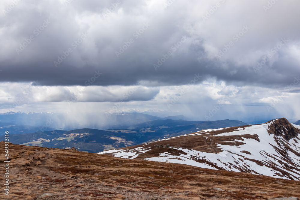 Panoramic view on summit cross of mountain peak Gertrusk seen from Ladinger Spitz, Saualpe, Carinthia, Austria, Europe. Snow covered hiking trail over alpine meadows and hills. Cloudy early spring day