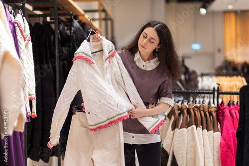 Second hand. Young Caucasian woman chooses sweater in store. In background there are d hangers with clothes. Concept of shopping and purchase © _KUBE_