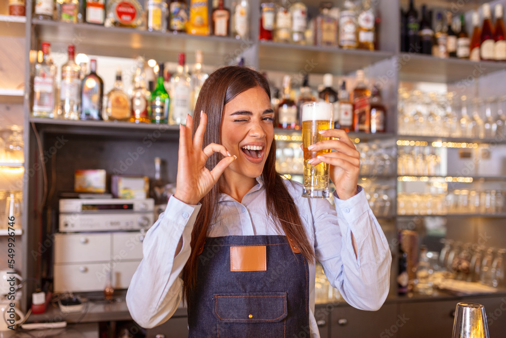 Beautiful smiling female Bartender showing ok sign serving a draft beer at the bar counter , shelves full of bottles with alcohol on the background Beautiful smiling female Bartender showing ok sign serving a draft beer at the bar counter , shelves full of bottles with alcohol on the background