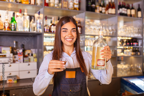 Beautiful female bartender is holding a shot glass with alcohol drink and a bottle in other hand, looking at camera and smiling while standing near the bar counter in cafe