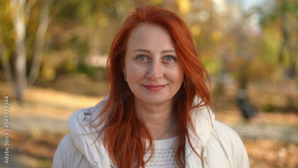 Front view portrait of mature Caucasian confident woman posing in sunny autumn park. Happy redhead lady smiling looking at camera standing outdoors Front view portrait of mature Caucasian confident woman posing in sunny autumn park. Happy redhead lady smiling looking at camera standing outdoors