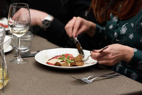 white dish with carpaccio of beef. a woman eats a small portion of carpaccio in a restaurant