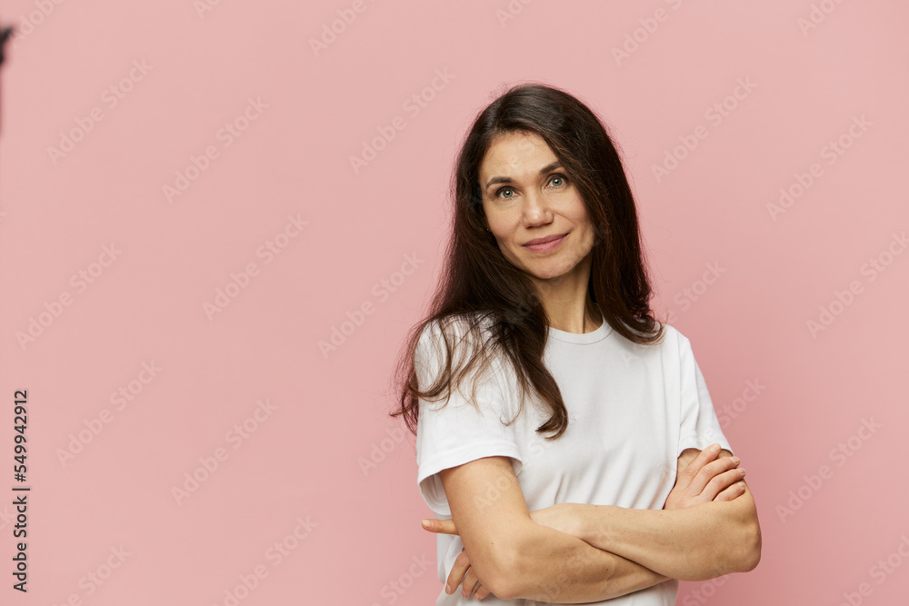 Fototapeta premium horizontal portrait of a cute, attractive woman on a pink background in a clean white t-shirt smiling pleasantly at the camera and folding her arms on her chest