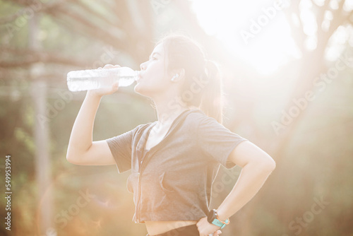 Fitness woman drinking water from sports bottle on afternoon workout after run training jogging outdoors at sunset. Girl wearing wireless headphones silhouette against sun flare.