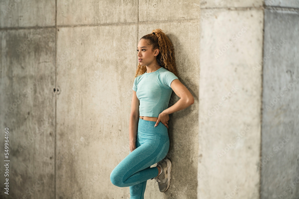 Young hispanic athletic woman leaning against a concrete wall recovering after physical exercise.