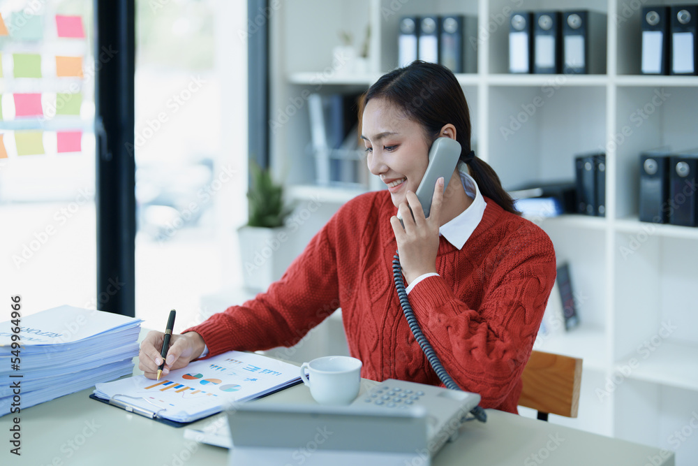 Portrait of a young Asian woman showing a smiling face as she uses her phone, computer and financial documents on her desk in the early morning hours Portrait of a young Asian woman showing a smiling face as she uses her phone, computer and financial documents on her desk in the early morning hours