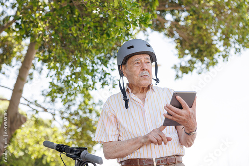 Fototapeta Naklejka Na Ścianę i Meble -  uomo anziano con caschetto di protezione utilizza il suo tablet per navigare in internet seduto vicino al proprio monopattino elettrico in un parco 