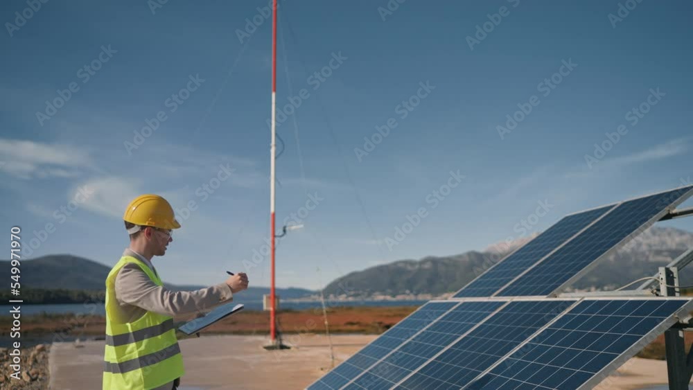 Male young inspector in protective vest and yellow helmet check the solar panel. Man engineer builder inspect weather station efficiency making notes. Energetics, engineering modern technology concept Male young inspector in protective vest and yellow helmet check the solar panel. Man engineer builder inspect weather station efficiency making notes. Energetics, engineering modern technology concept