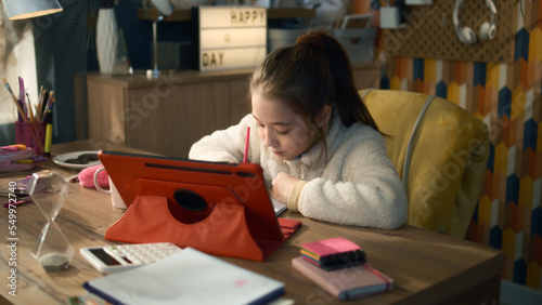 Young asian girl sits at the table at cozy room and talks with teacher. Schoolgirl listens online lesson, studies on tablet,writes down and discuss new material. Distance learning concept.