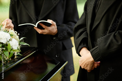 Unrecognizable woman wearing black at outdoor funeral ceremony with priest reading prayer in background  copy space