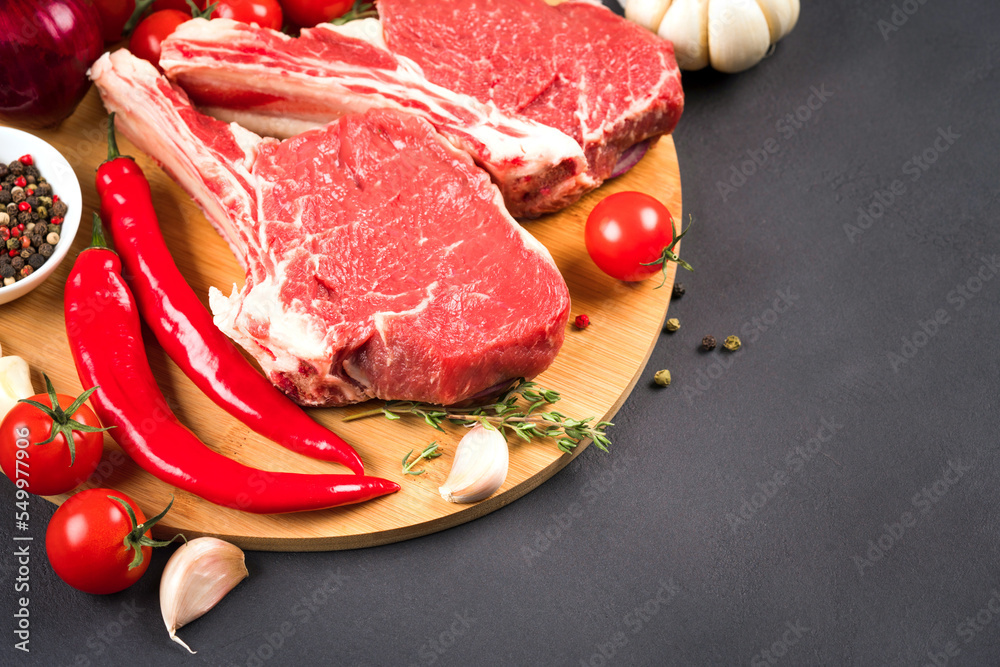 Cowboy steak recipe. Raw ribeye beef steak ready for grilling with seasonings and chili pepper on wooden cutting board over dark concrete background with copy space for text Cowboy steak recipe. Raw ribeye beef steak ready for grilling with seasonings and chili pepper on wooden cutting board over dark concrete background with copy space for text