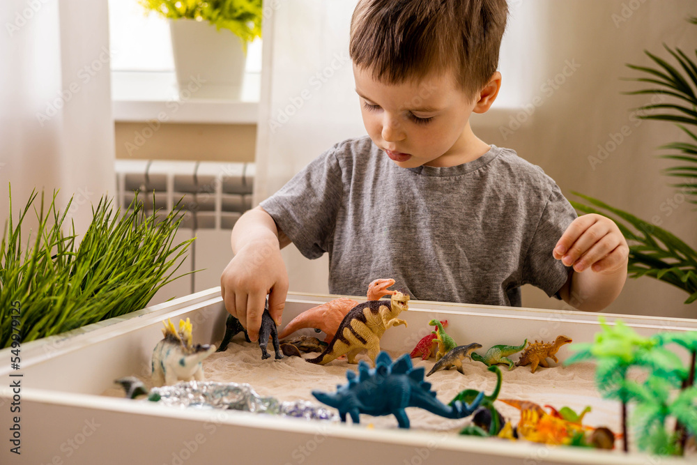Cute baby boy playing sensory box kinetic sand table with carnivorous and herbivorous dinosaurs - obrazy, fototapety, plakaty 
