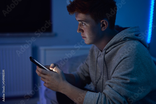 Thoughtful caucasian teenage boy using mobile phone while sitting at night in his room and looking away