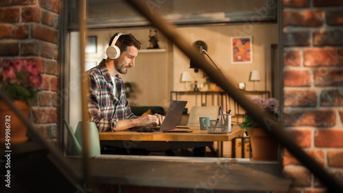 Handsome Caucasian Man Working on Laptop Computer, Sitting Behind His Desk at Home Office. Freelance Entrepreneur Listening to Music, Doing Remote Telework. View From Outdoors into Window.