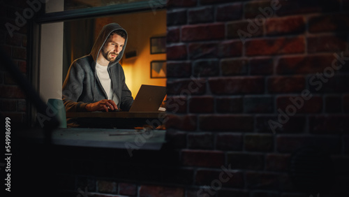 Information Technology Specialist Working Late on a Latptop. Software Engineer Wearing Hoodie, Working on Computer in His Cozy Home Office Apartment. Remote Work. Night View From Outdoors into Window. photo