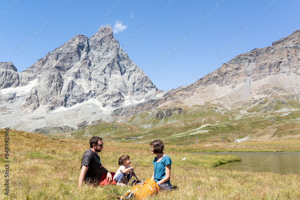 Fototapeta premium Happy family with toddler sitting in a green meadow near a lake with views at Matterhorn at italian alps.