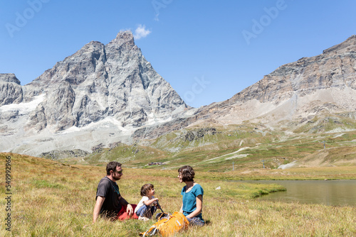 Happy family with toddler sitting in a green meadow near a lake with views at Matterhorn at italian alps.