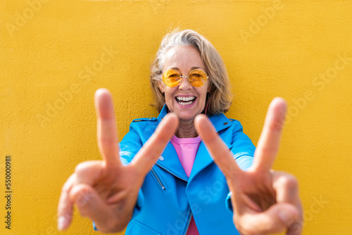 Happy mature woman gesturing peace sign in front on yellow wall