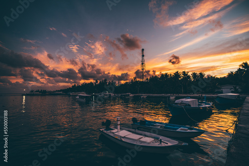 Wide-angle low-key view of a jetty on a tropical tourist island with an atmospheric sunset in the background; an ocean pier with a few speedboats and ferry vessels with a dramatic evening cloudscape