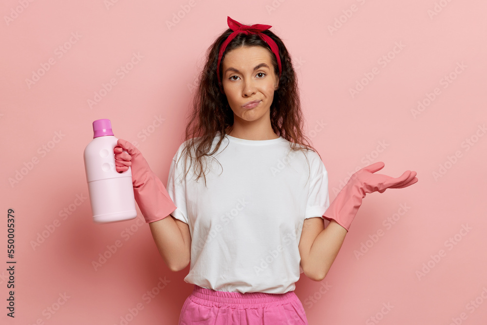Young bored housewife, stands isolated over pink wall, holds sanitising liquid with upset face emotion Young bored housewife, stands isolated over pink wall, holds sanitising liquid with upset face emotion