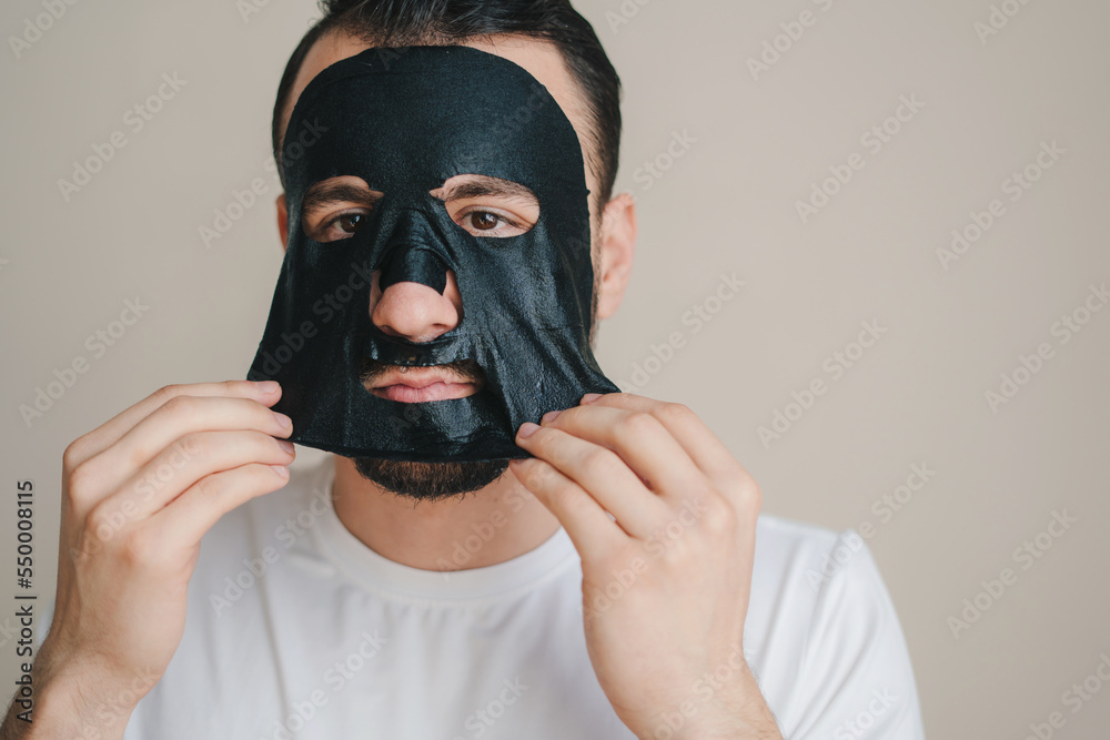 Portrait of a young man peeling off a black facial mask isolated over gray background. Health care. Beauty face. Beauty portrait. Beauty treatment. Aging care. Portrait of a young man peeling off a black facial mask isolated over gray background. Health care. Beauty face. Beauty portrait. Beauty treatment. Aging care.