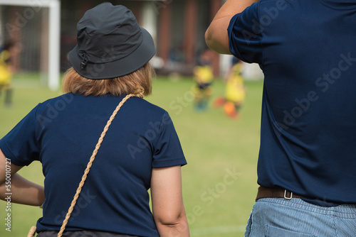 Mom and dad standing and taking pictures their son playing football in a school tournament on a sideline with a sunny day. Sport, outdoor active, lifestyle and happy family.