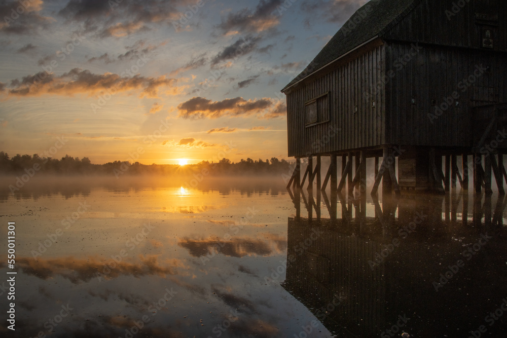 Pfahlhaus am Teich bei Sonnenaufgang. Nebel über und Spiegelung im Wasser. Morgenstimmung