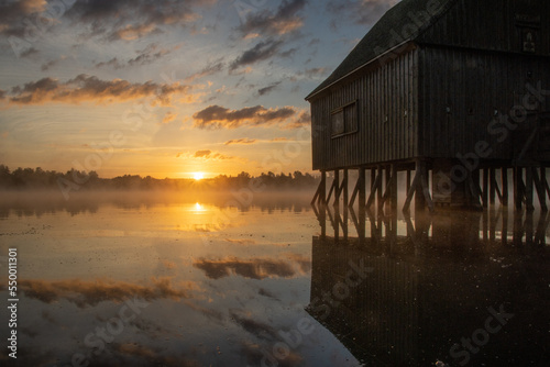 Pfahlhaus am Teich bei Sonnenaufgang. Nebel über und Spiegelung im Wasser. Morgenstimmung