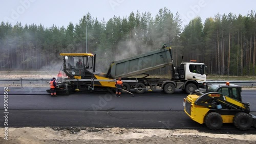 Road works. Laying new asphalt on an intercity highway. The truck pours the hot mix into the paver. Workers in overalls are watching the process. In the foreground is a small excavator. Roadbed repair