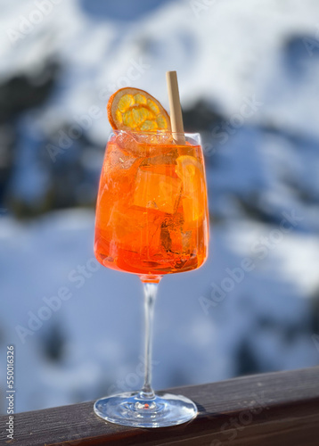 A glass with an orange alcoholic cocktail against the backdrop of snow-capped mountains. Freshly made aperol spritz in outdoor cafe. The atmosphere of a cool winter vacation in a ski resort. Nobody.