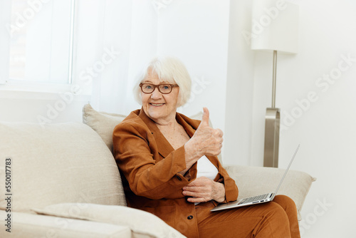 a broadly smiling elderly woman enjoys career success sitting on the couch, working from home on a laptop, giving a thumbs up © Tatiana