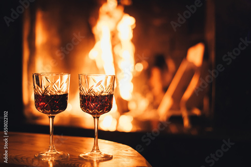Two glasses of red wine on wooden table with cozy fireplace flame on the background.
