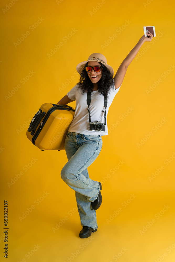 Happy Traveller curly latin woman wears white t-shirt with suitcase bag, passport and boarding tickets isolated on yellow background. Happy Traveller curly latin woman wears white t-shirt with suitcase bag, passport and boarding tickets isolated on yellow background.
